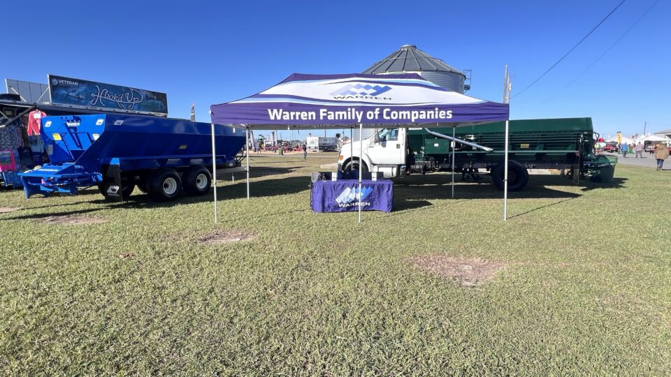 Warren Family of Companies canopy between blue pull-type and green truck-mounted spreaders at Sunbelt Ag Expo